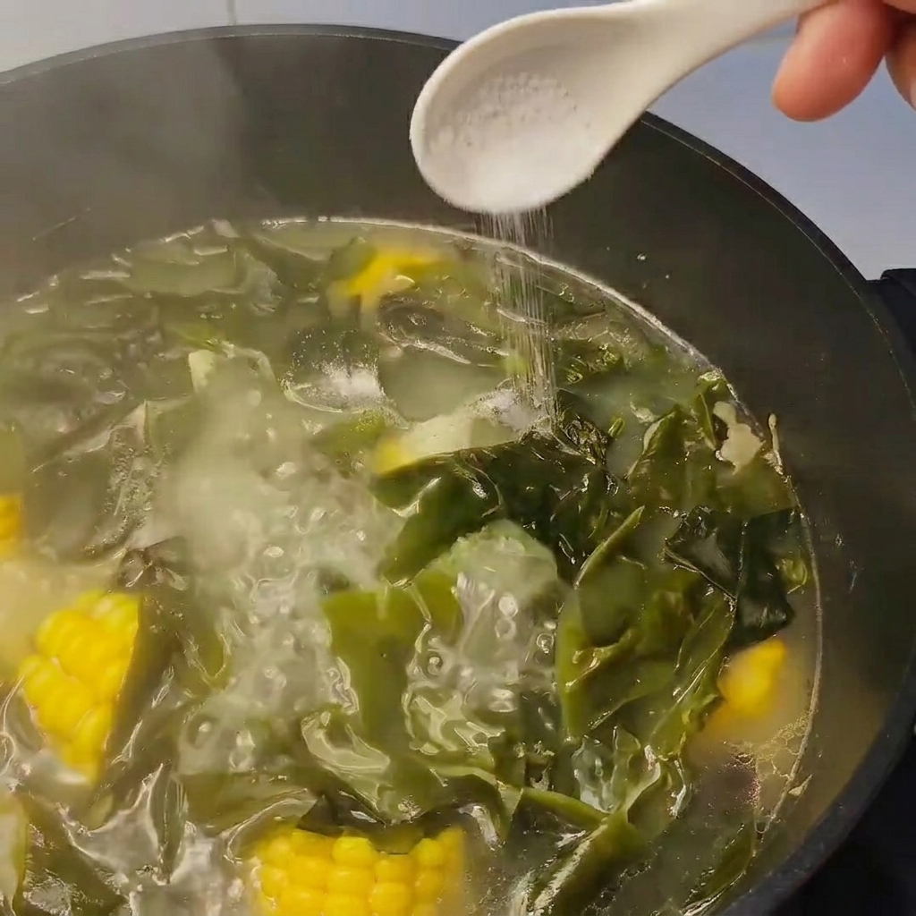 Una cucharada de sal siendo añadida a una olla de sopa a fuego lento con alga y maíz.