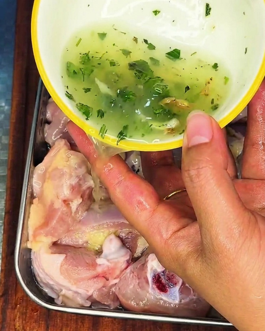 Una mano vertiendo un líquido con trozos de cebolleta y jengibre sobre el pollo crudo en un recipiente de acero inoxidable.