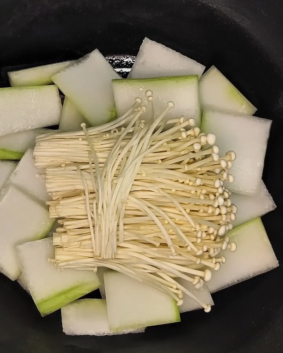 Un manojo de champiñones enoki extendidos sobre la capa de rodajas de melón de invierno en la olla.