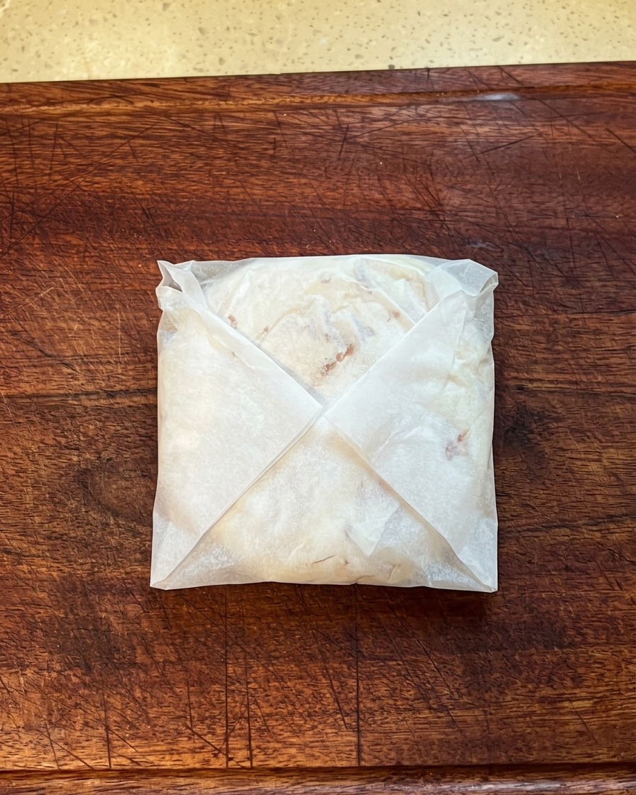 Un panqueque de cebolleta doblado y envuelto en papel de horno, presionándose para aplanarlo sobre una tabla de cortar de madera.