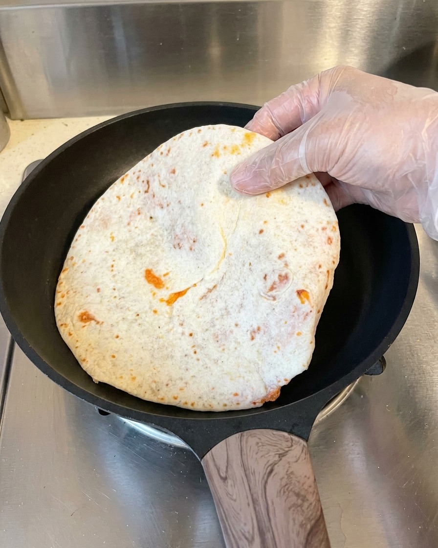 Una mano con un guante de plástico coloca un panqueque aplanado y envuelto en papel de horno en una sartén antiadherente negra y caliente.