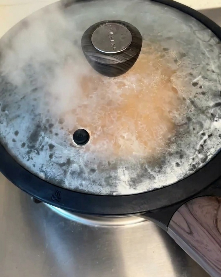 Una sartén antiadherente tapada sobre los fogones, con vapor visible escapando por debajo de la tapa mientras se cocina el panqueque.
