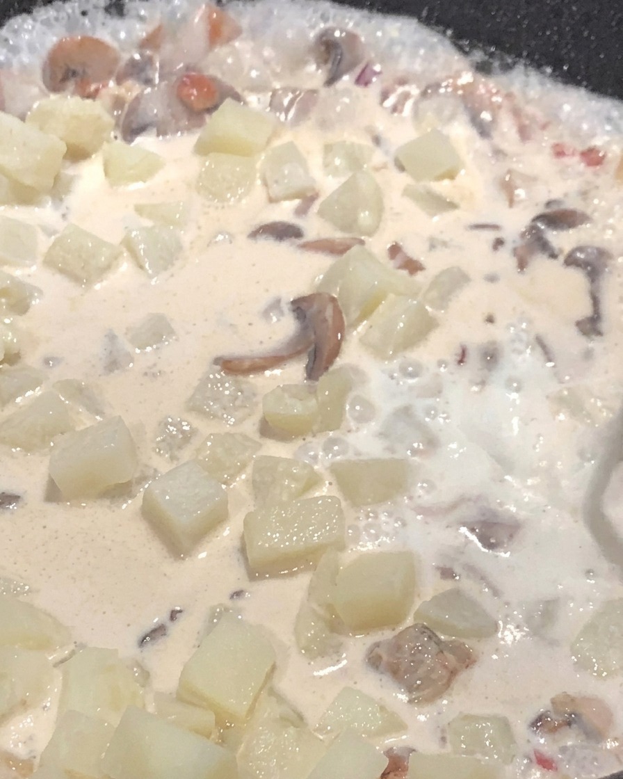 Leche siendo vertida en una sartén llena de papas y champiñones, hirviendo a fuego lento en una salsa cremosa.