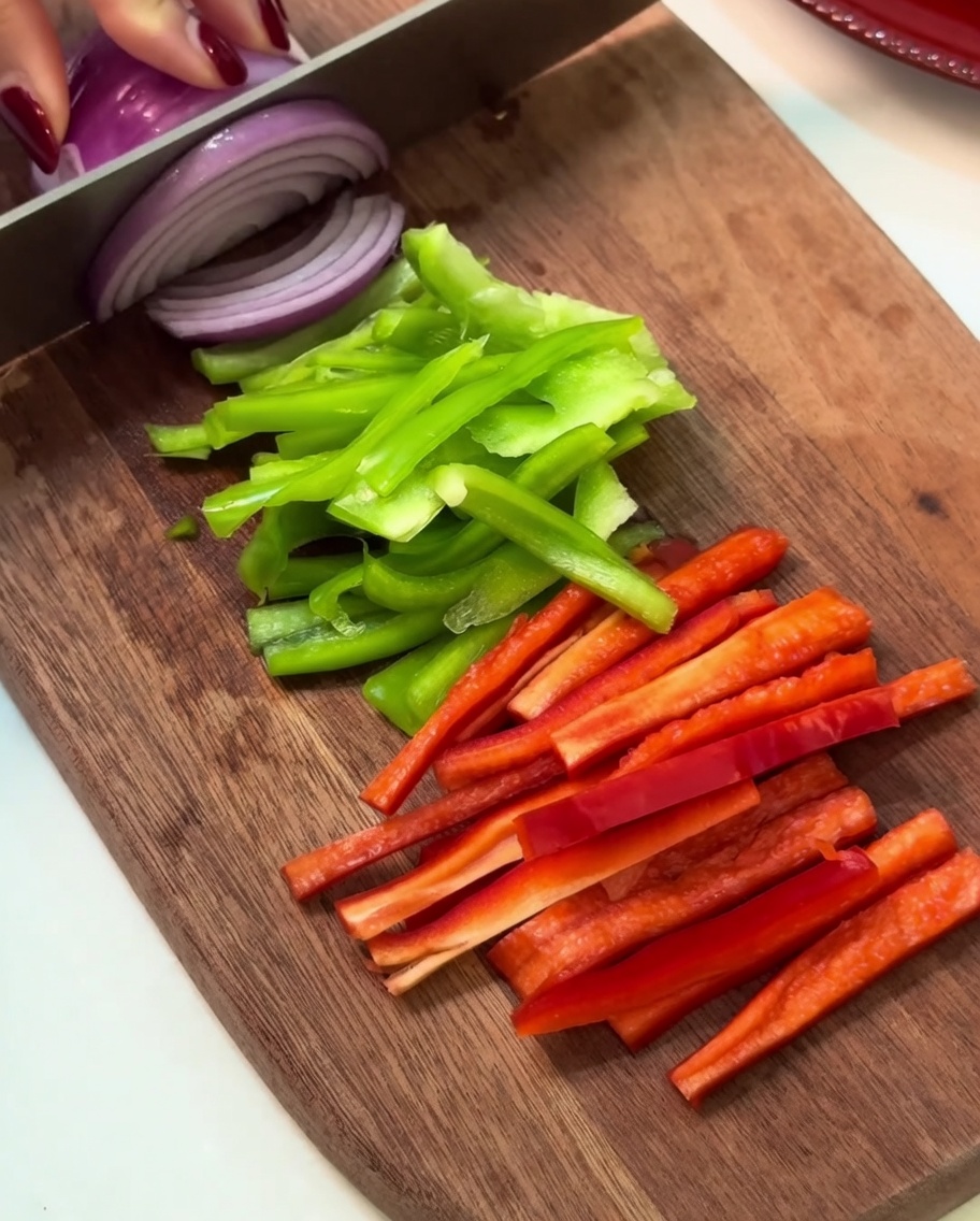 Tiras de pimiento verde, pimiento rojo y cebolla morada dispuestas ordenadamente sobre una tabla de cortar de madera.