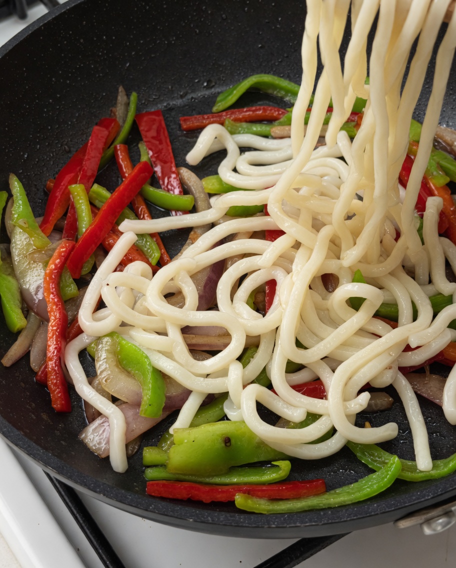 Fideos udon y tiras de ternera siendo añadidos a la sartén con las verduras.