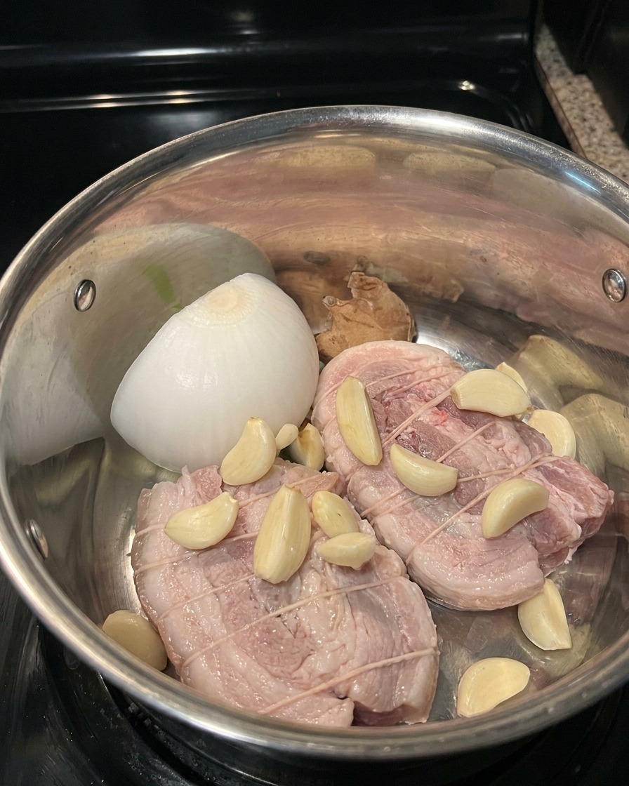 Trozo de panceta de cerdo con aromáticos y líquido de braseado en una olla de acero inoxidable.