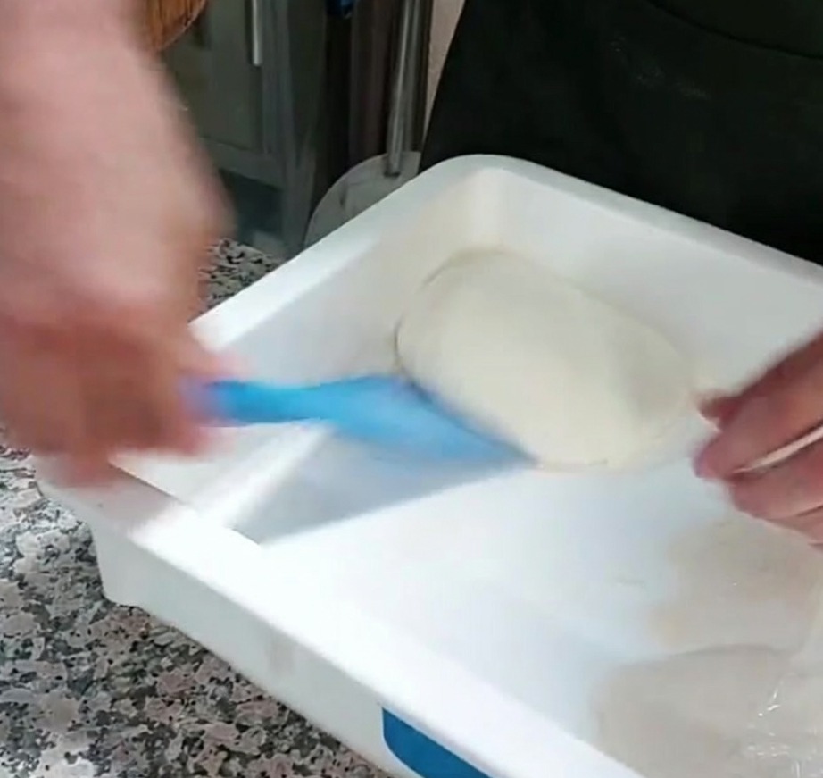 Una porción de masa de pizza siendo levantada de un recipiente usando una espátula de plástico azul.