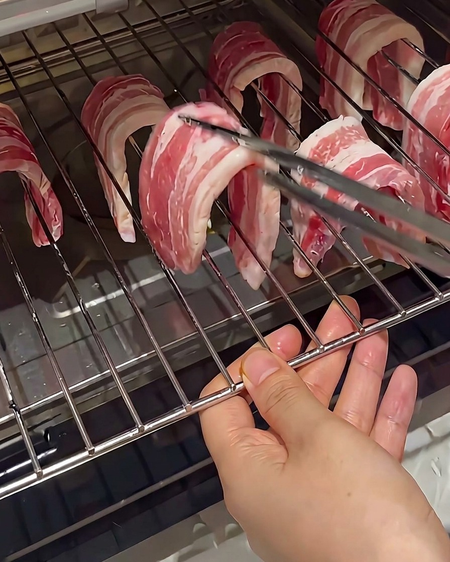 Tiras de panceta cruda colgadas sobre los alambres de una rejilla de horno, sujetas con pinzas.