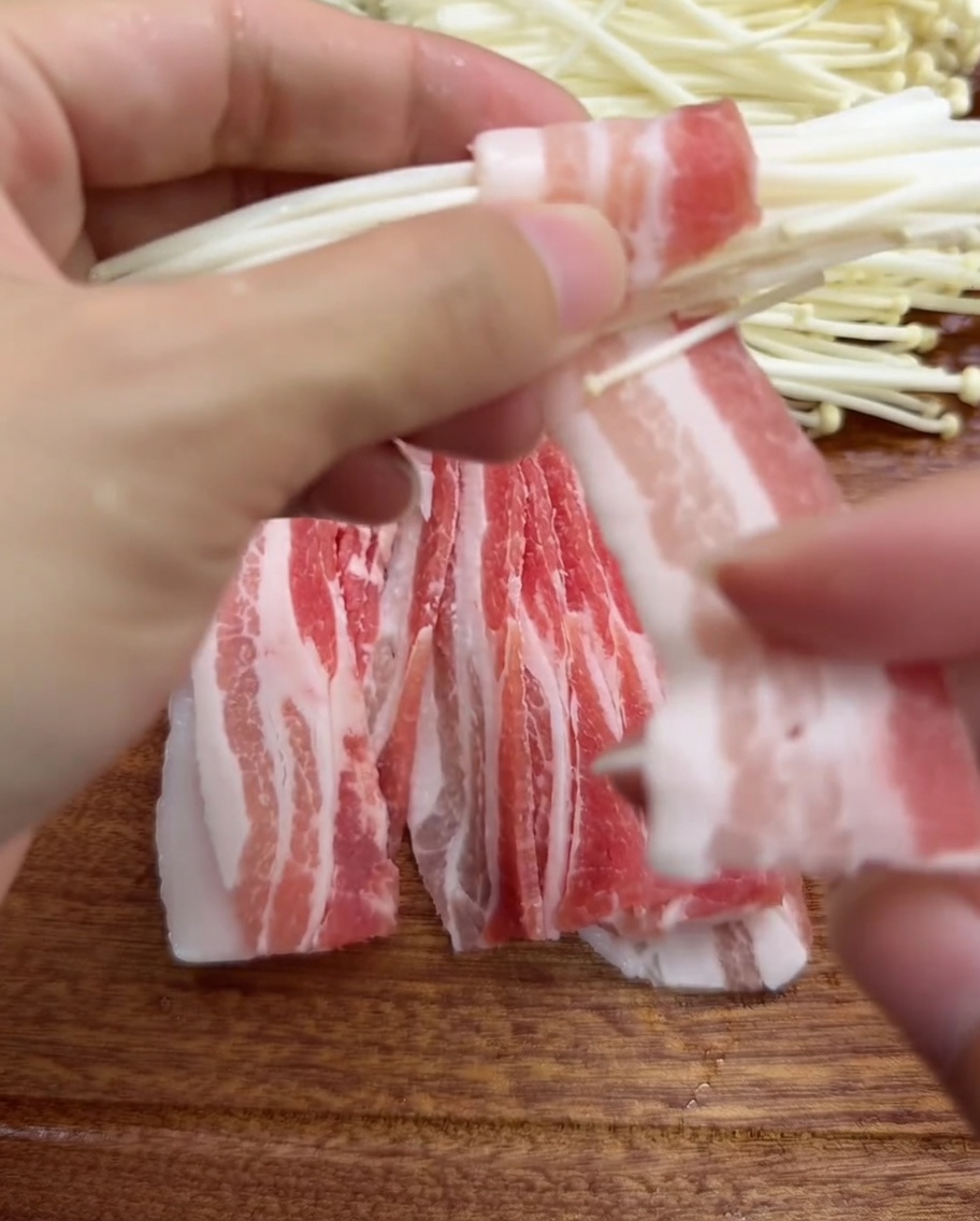 Primer plano de unas manos envolviendo una fina rebanada de panceta de cerdo alrededor de un racimo de setas enoki.