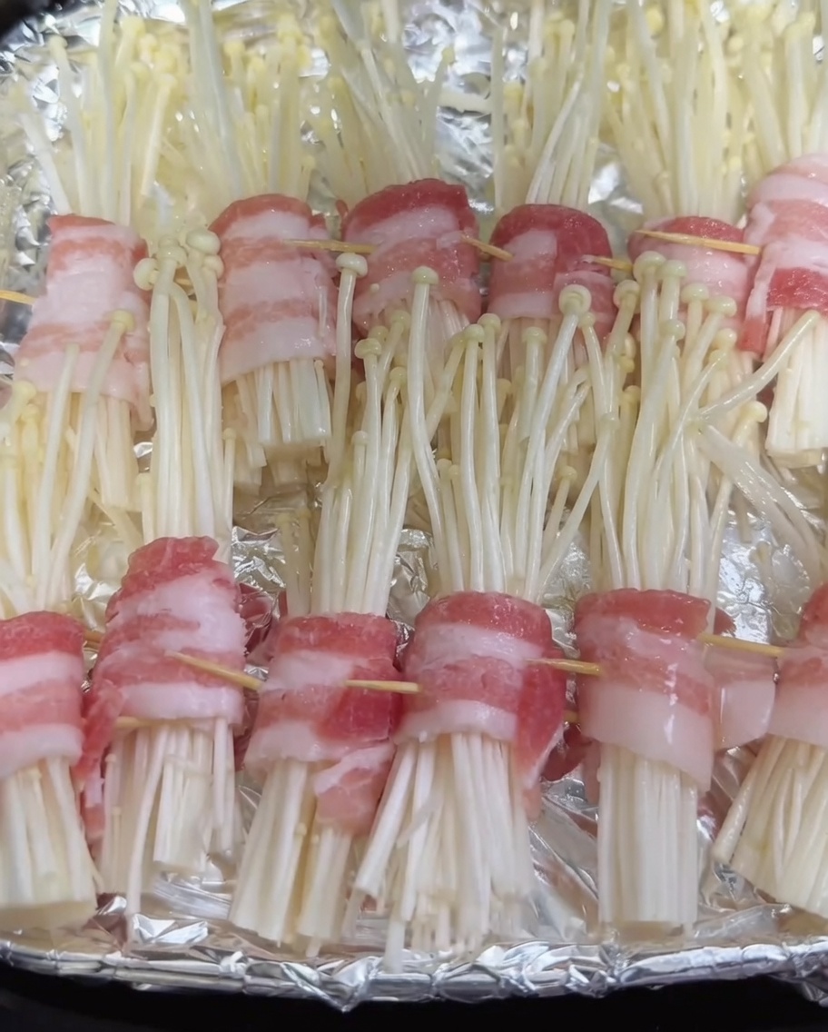 Botella pulverizadora aplicando una fina capa de aceite sobre los rollitos crudos de panceta y enoki en la freidora de aire.