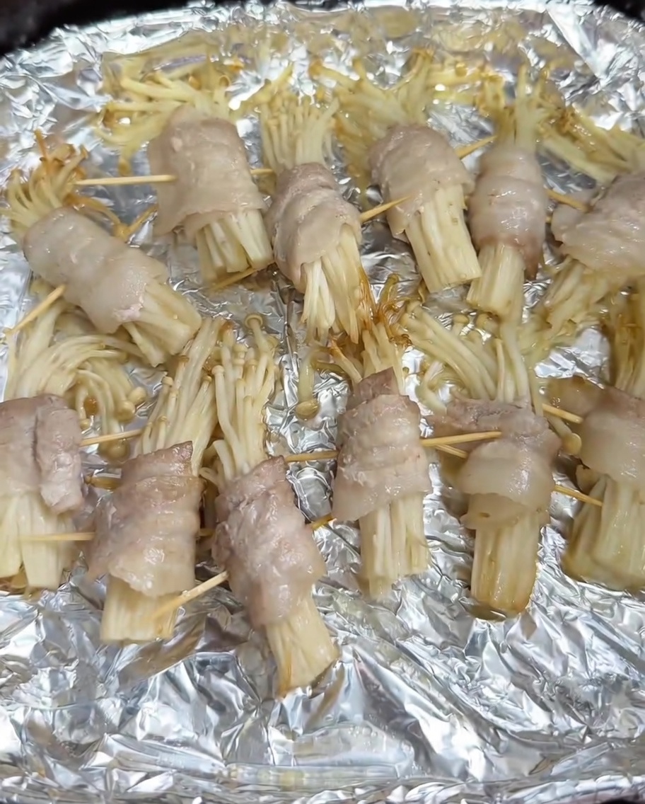 Sazonador de barbacoa espolvoreándose sobre los rollitos de panceta y enoki medio cocidos en la cesta de la freidora de aire.