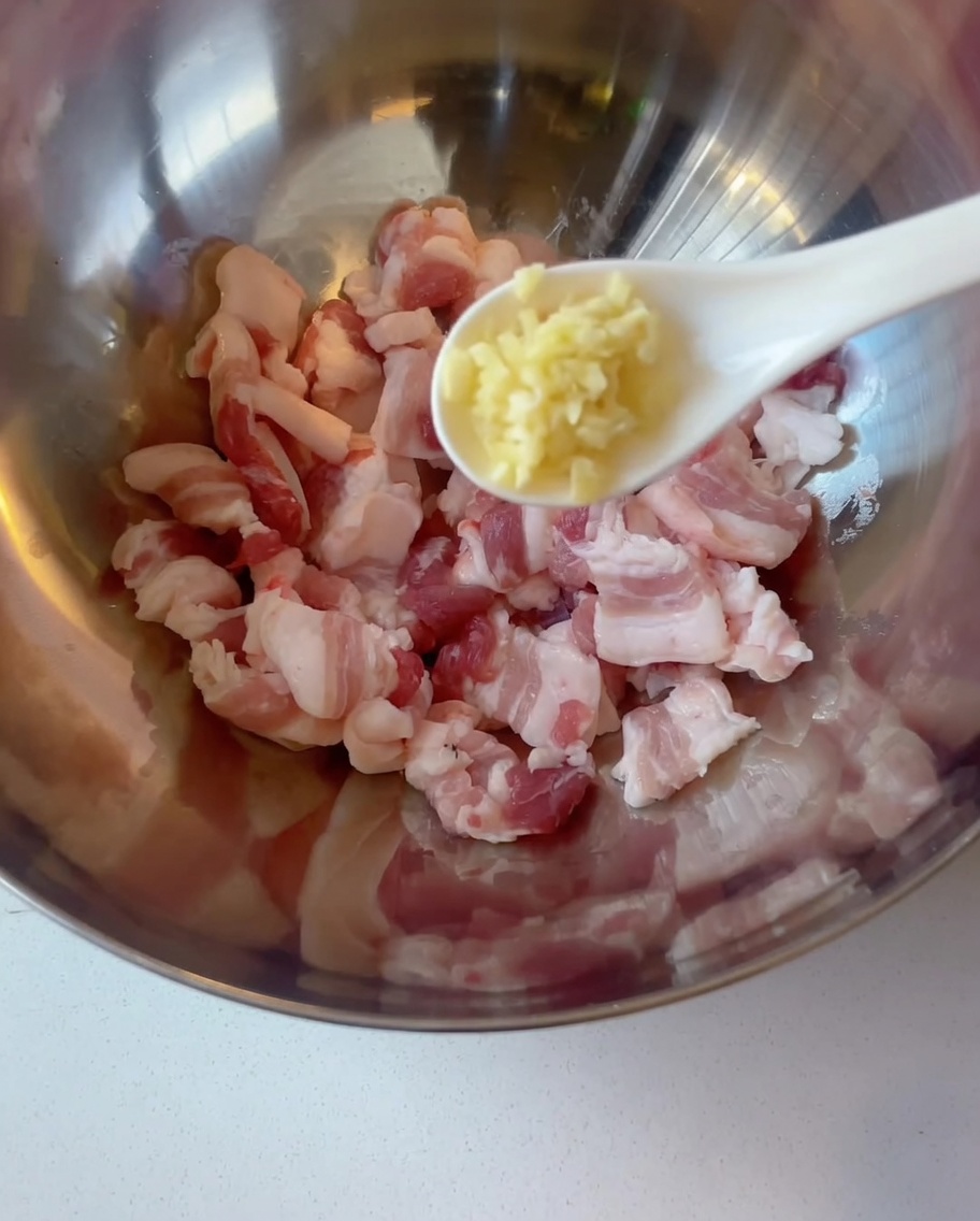 Rebanadas de panceta cruda en un tazón de metal mientras se añade ajo picado con una cuchara.