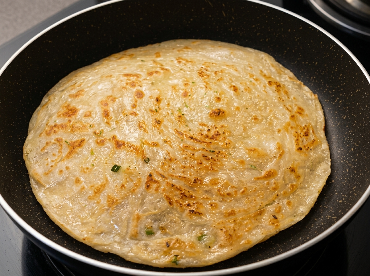 Un pancake de cebolleta redondo friéndose en una sartén antiadherente oscura, mostrando una superficie dorada y texturizada.