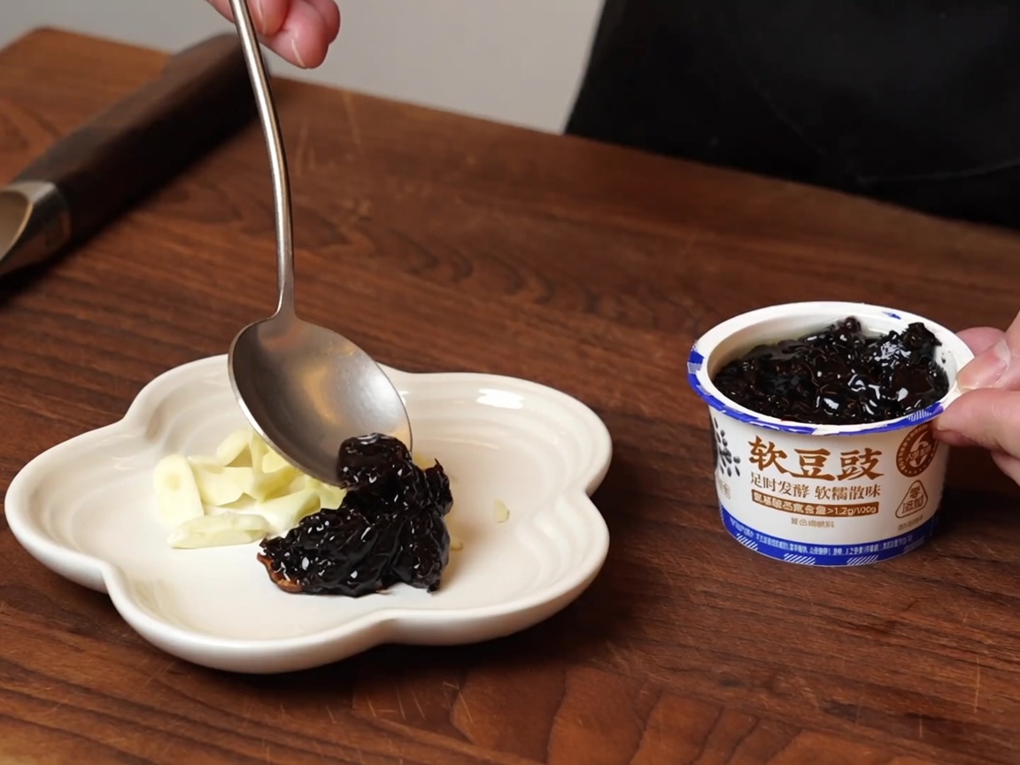 Una cuchara sirviendo frijoles negros fermentados húmedos en un plato pequeño junto al ajo.