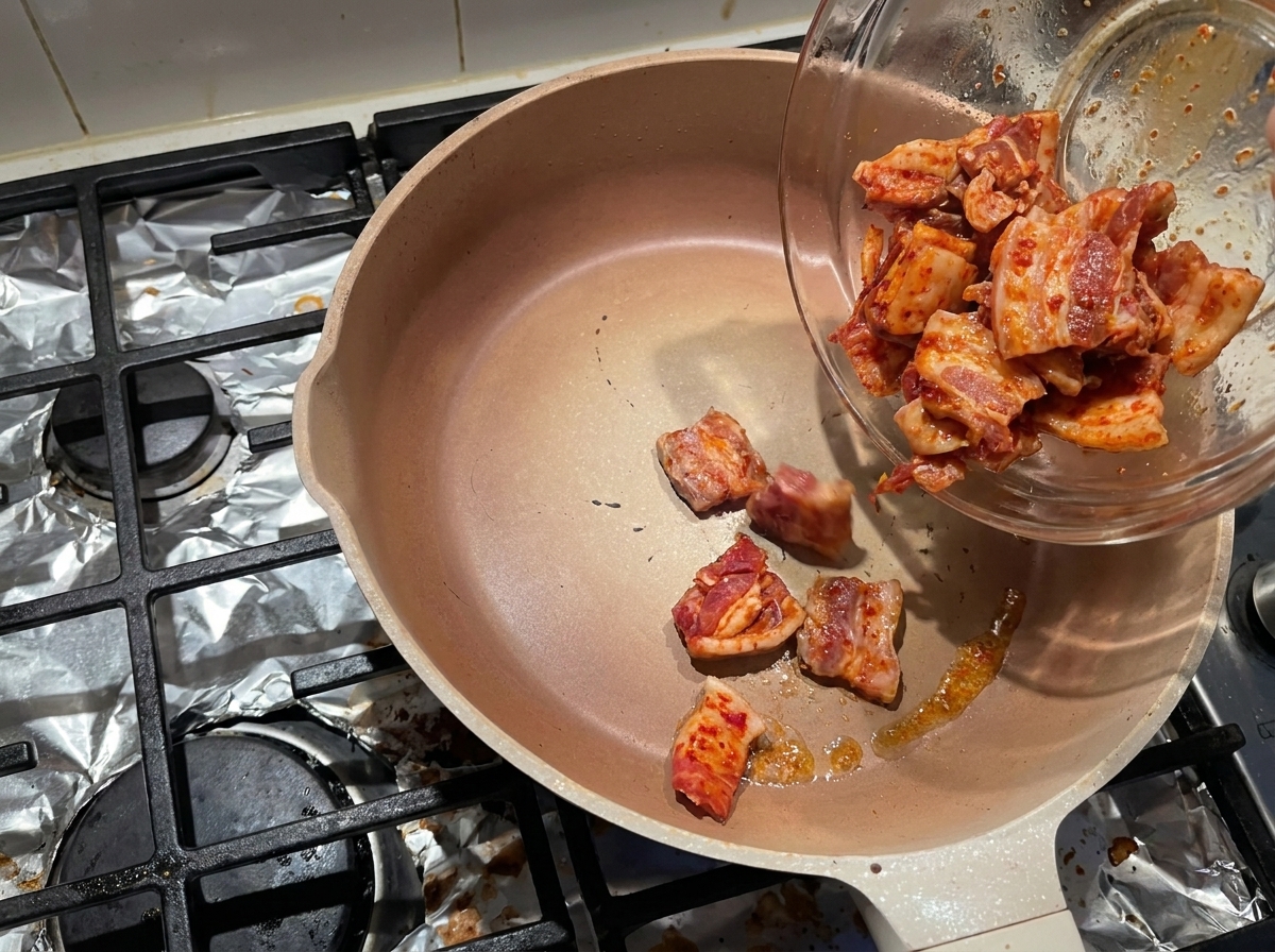 Láminas de panceta marinada deslizándose desde un bol hacia una sartén caliente sin aceite para renderizar la grasa.