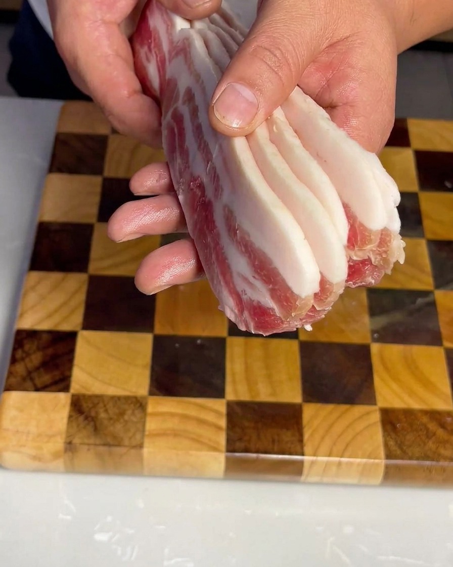 Tiras uniformes de panceta de cerdo cruda dispuestas cuidadosamente sobre una tabla de cortar de madera.