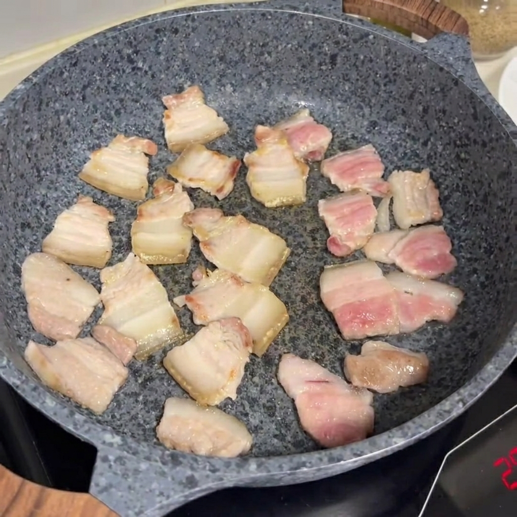 Rodajas de panceta friéndose y dorándose en una sartén de piedra gris.