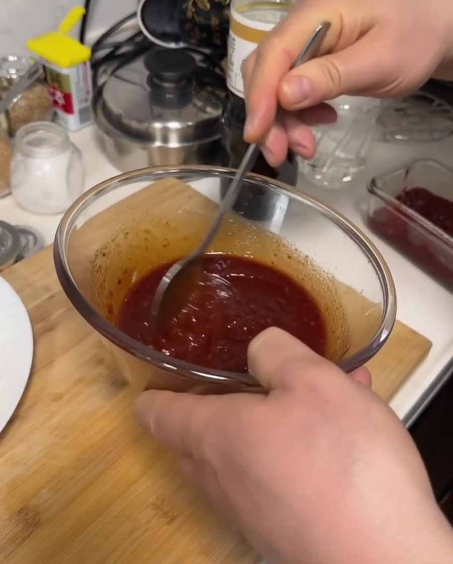 Una mano usando una cuchara de metal para mezclar una salsa roja oscura y espesa en un cuenco de cristal.