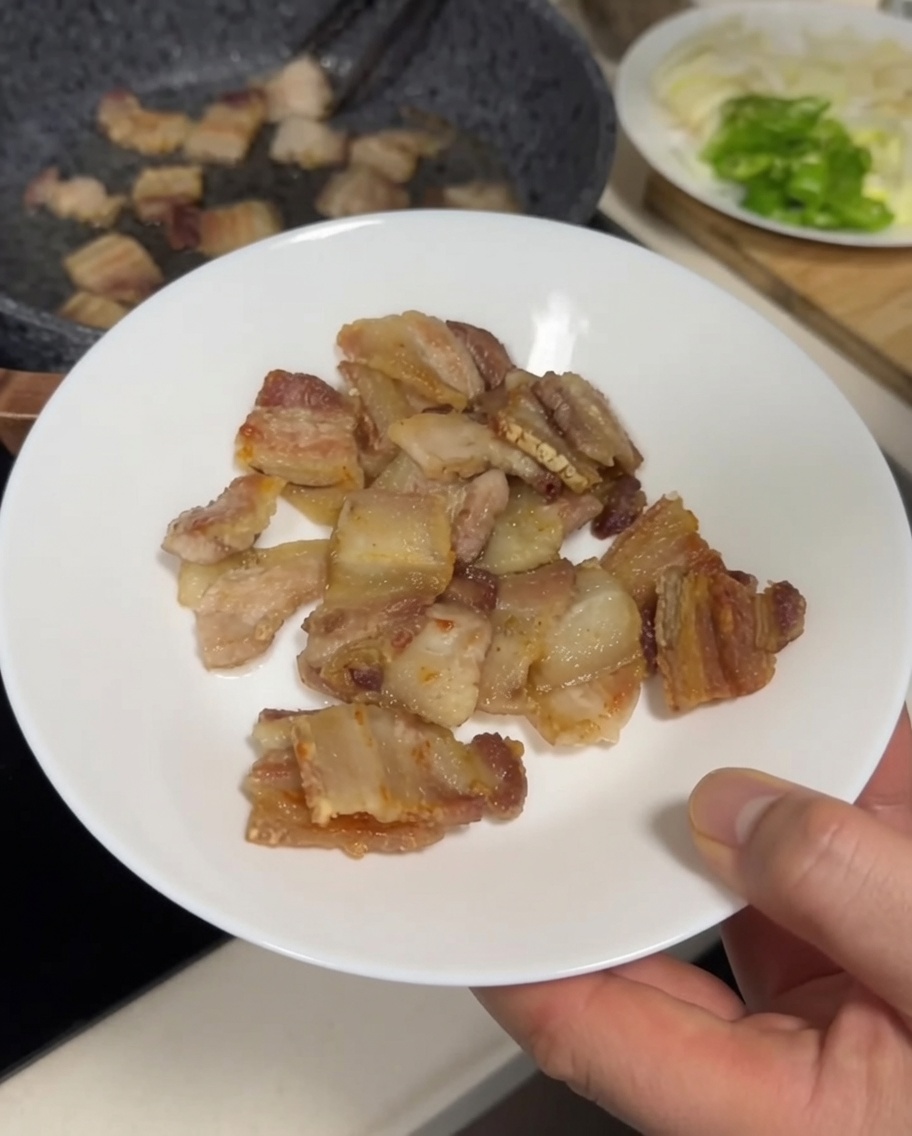 Trozos de panceta dorados y crujientes siendo transferidos de la sartén a un plato blanco con pinzas de metal.