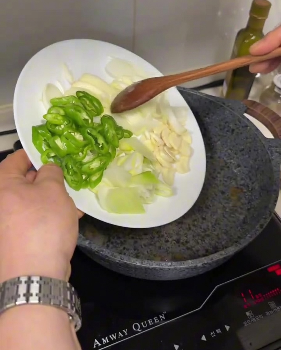 Chiles verdes en rodajas, cebollas blancas y ajo siendo añadidos desde un plato a una sartén caliente con aceite.