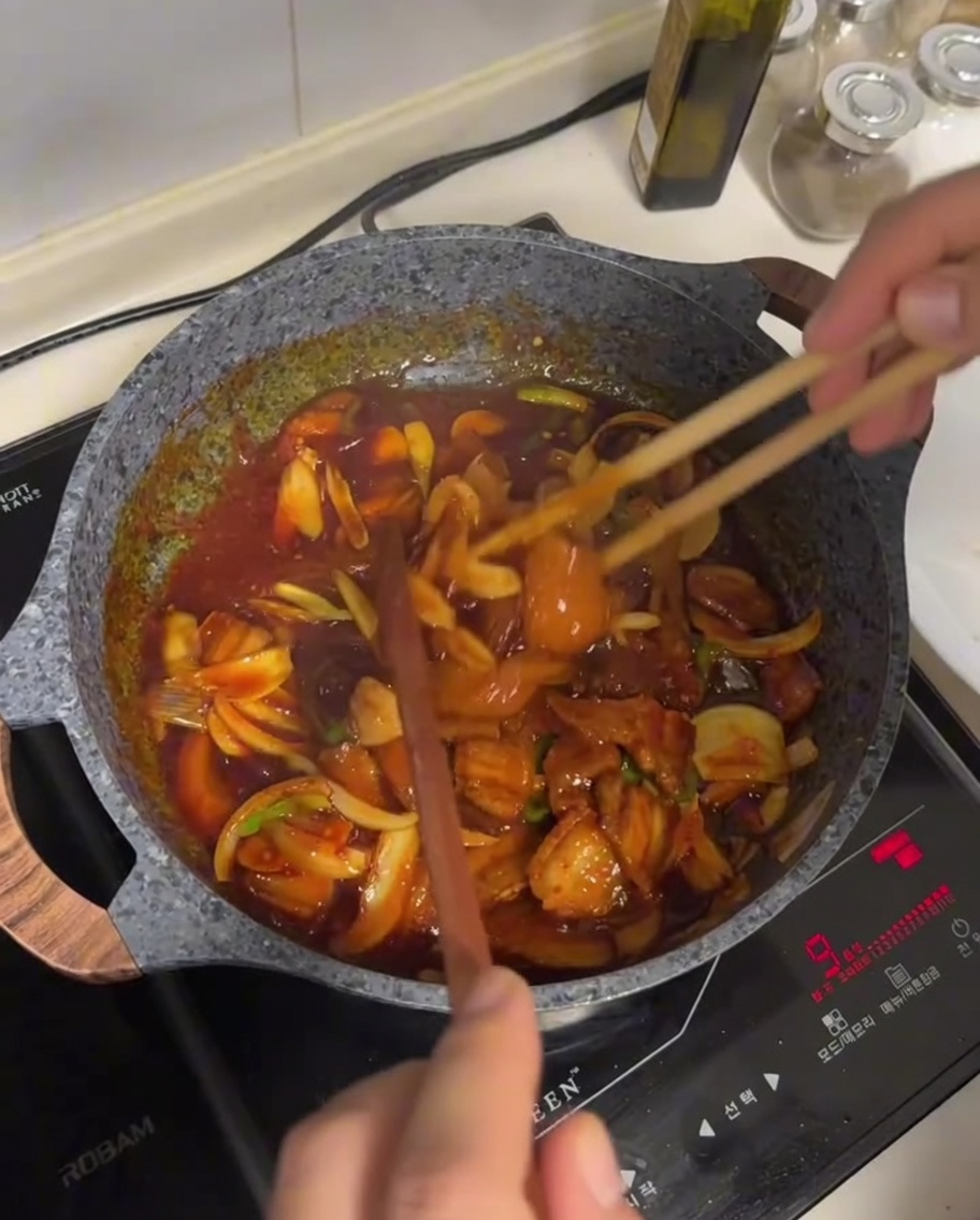 La mezcla de panceta y verduras siendo salteada en una salsa roja brillante y oscura usando palillos de madera largos en una sartén antiadherente.