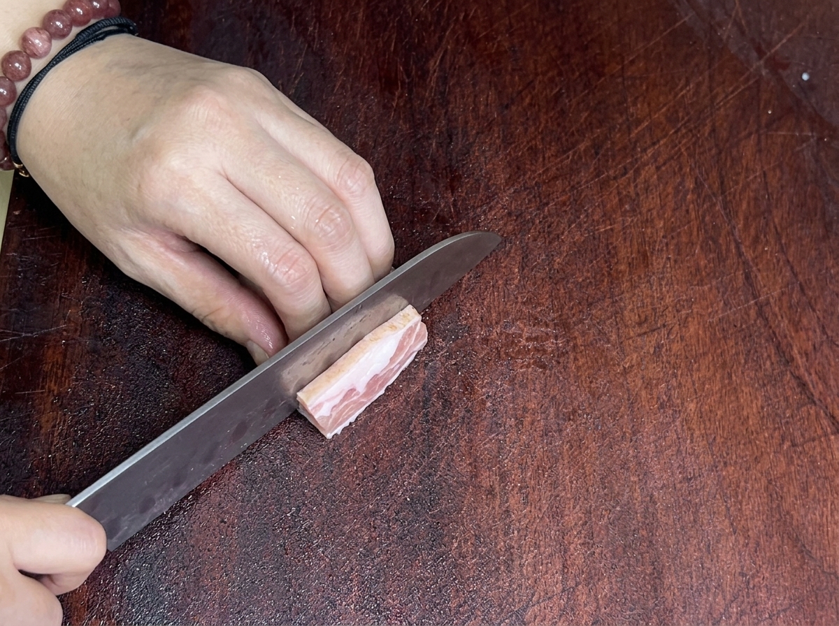 Primer plano de un cuchillo afilado cortando una pieza de panceta sobre una tabla de madera.