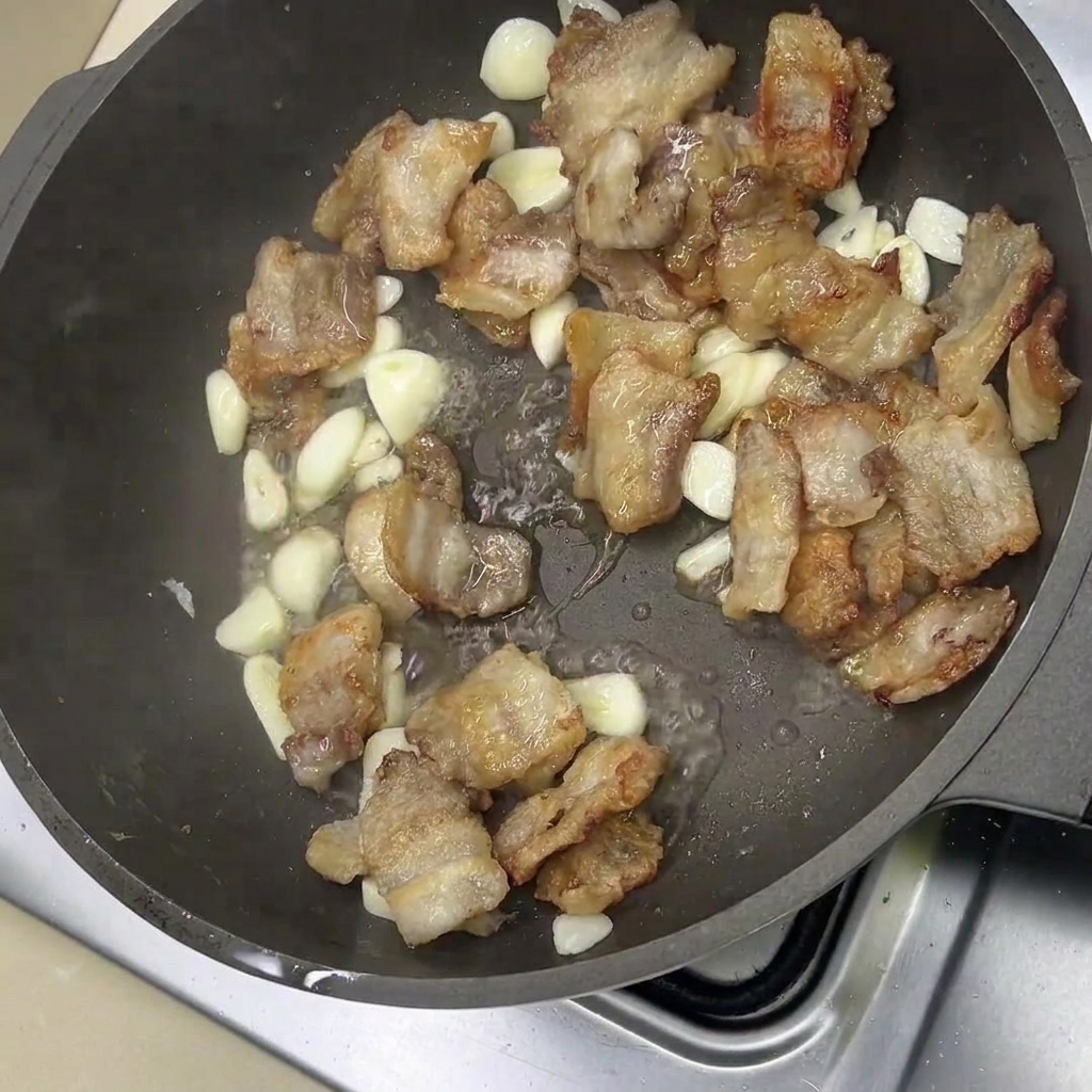 Rodajas de panceta frita y dientes de ajo enteros chisporroteando en la sartén.