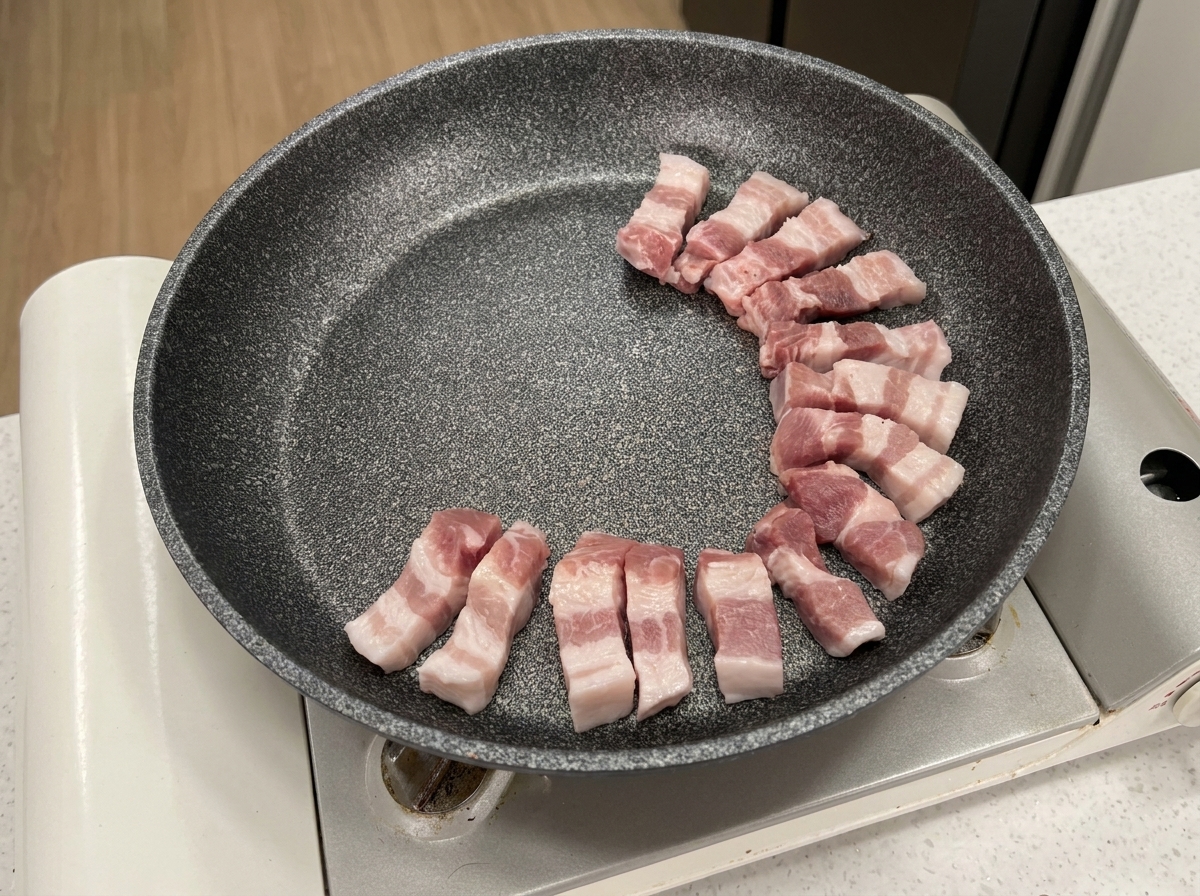 Tiras de panceta de cerdo cruda dispuestas en círculo en una sartén antiadherente gris.