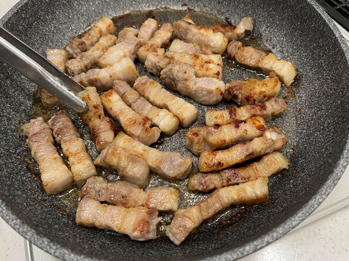 Trozos de panceta de cerdo volteados con pinzas en una sartén, mostrando superficies doradas.