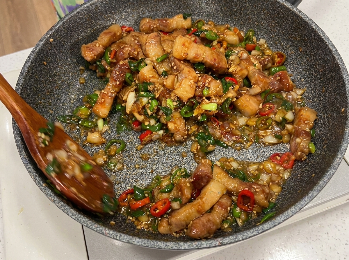 Trozos de panceta de cerdo, chile y cebollino mezclados en la sartén con dos cucharas de madera.