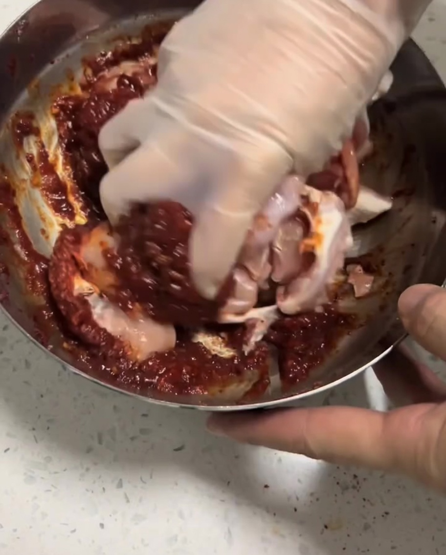 Trozos de pollo crudo siendo masajeados por una mano con guante en la marinada roja dentro de un tazón.