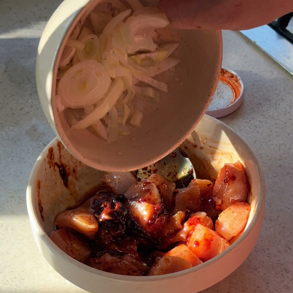 Cebolla en tiras y ajo picado añadiéndose a un bol con trozos de pollo condimentados.