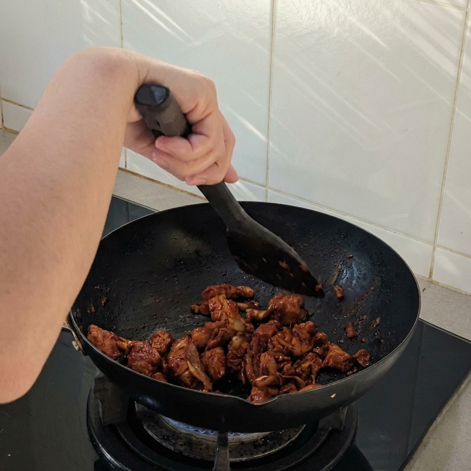 Trozos de pollo y cebolla salteándose en un wok negro con una espátula, cubiertos de una salsa picante oscura.