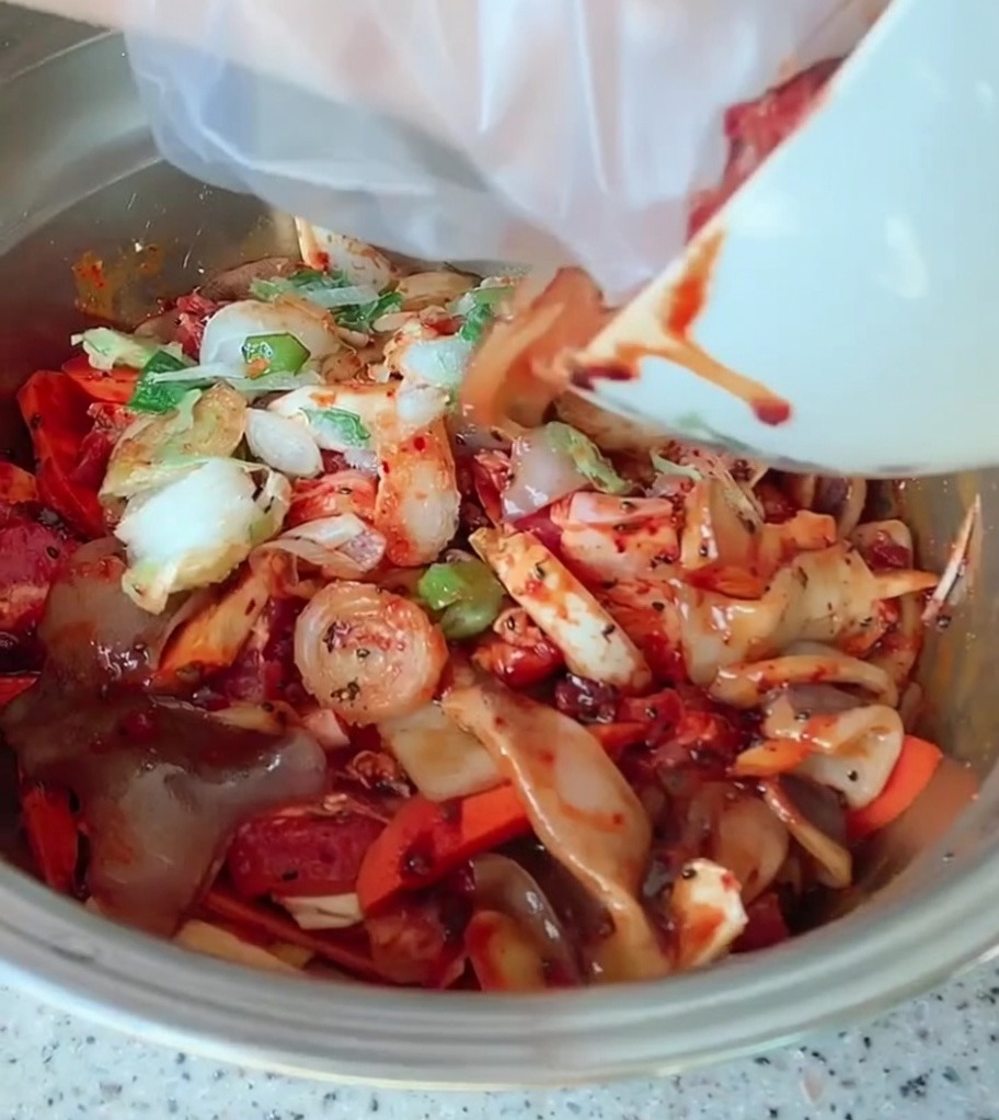 Un gran tazón de metal con rodajas de panceta de cerdo, verduras coloridas y cebolleta cubiertos de una vibrante salsa roja picante.