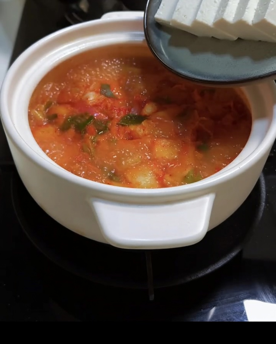 Tofu en rodajas siendo añadido a la hirviente sopa roja de kimchi.