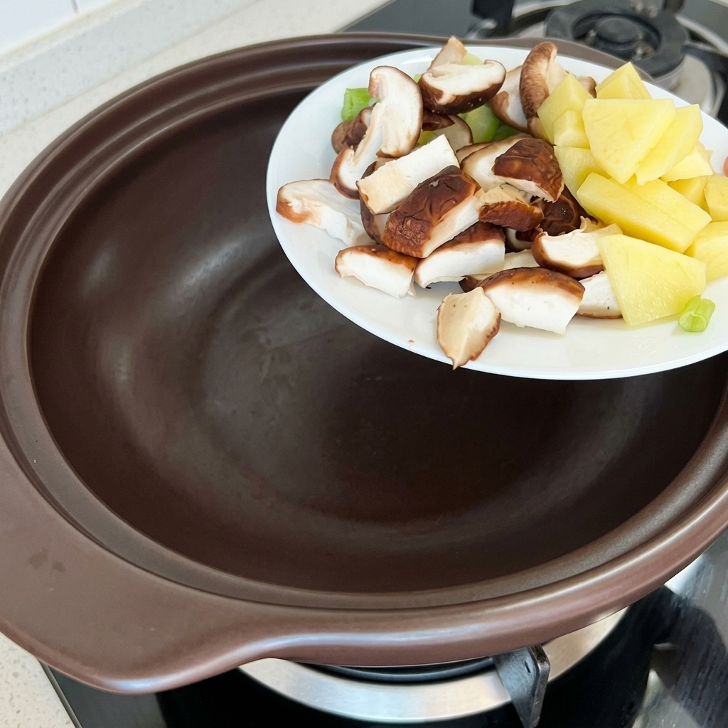 Patatas, calabacín y setas shiitake picadas siendo añadidas a una cazuela de barro.