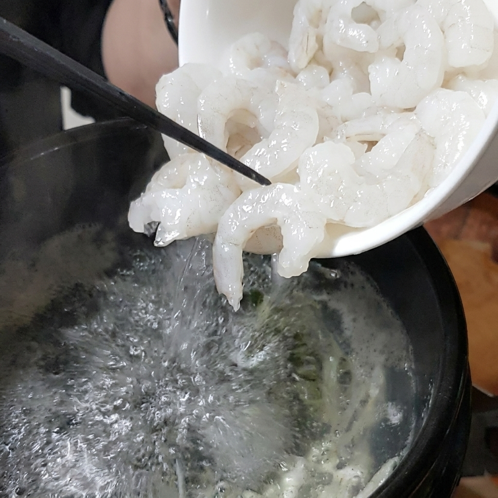 Un tazón blanco vertiendo camarones frescos en una olla de piedra burbujeante con caldo.
