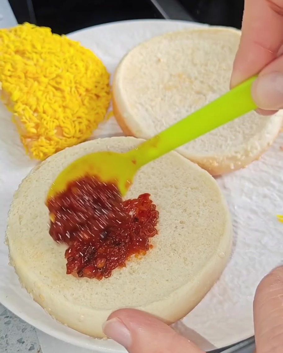 Espátula pequeña extendiendo salsa picante sobre el pan de hamburguesa tostado.
