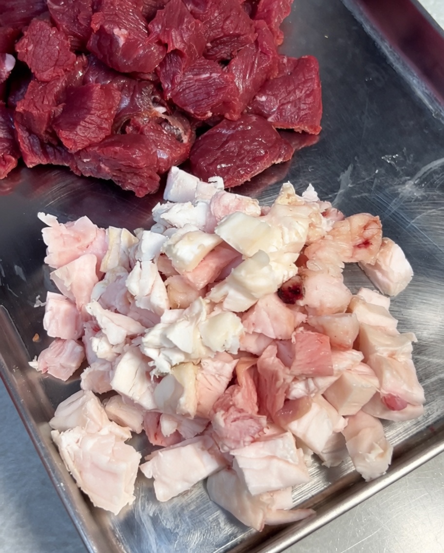 Trozos de carne y grasa siendo sazonados con sal y pimienta negra en un bol de acero inoxidable.