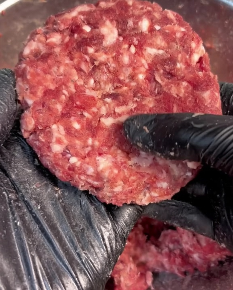 Mano con guante negro dando forma de disco a carne picada sazonada.
