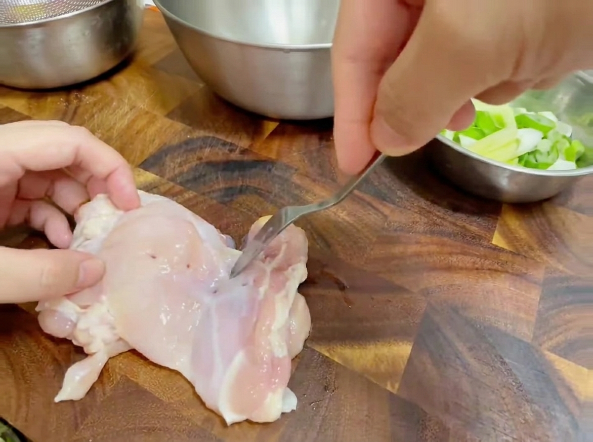 Primer plano de un tenedor pinchando un muslo de pollo crudo deshuesado sobre una tabla de cortar de madera.
