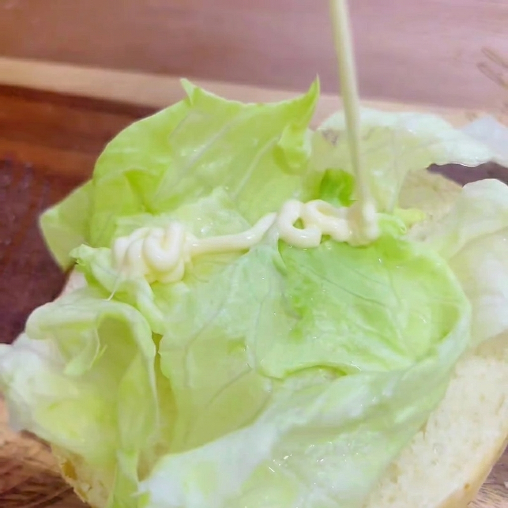 Mayonesa blanca siendo vertida en forma de zigzag sobre la lechuga fresca en el pan de hamburguesa.