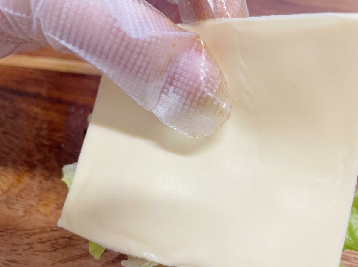Una mano con guante colocando cuidadosamente una loncha de queso blanco sobre la hamburguesa en montaje.