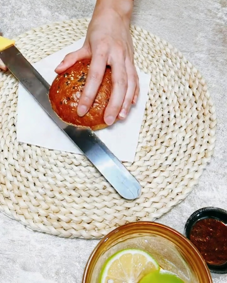 Una mano sosteniendo un cuchillo mientras corta un pan de hamburguesa artesanal por la mitad.
