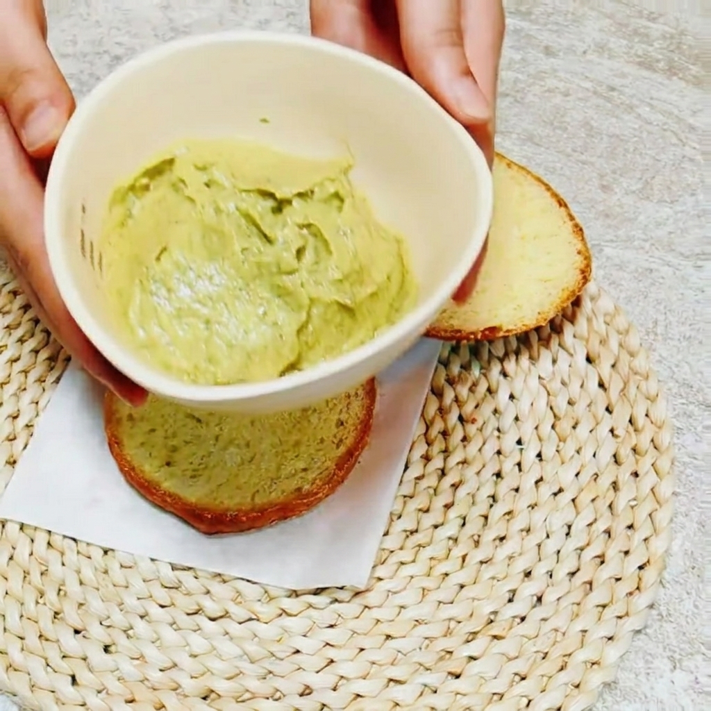 Primer plano de un cuchillo extendiendo salsa de aguacate y albahaca sobre el interior de un pan.