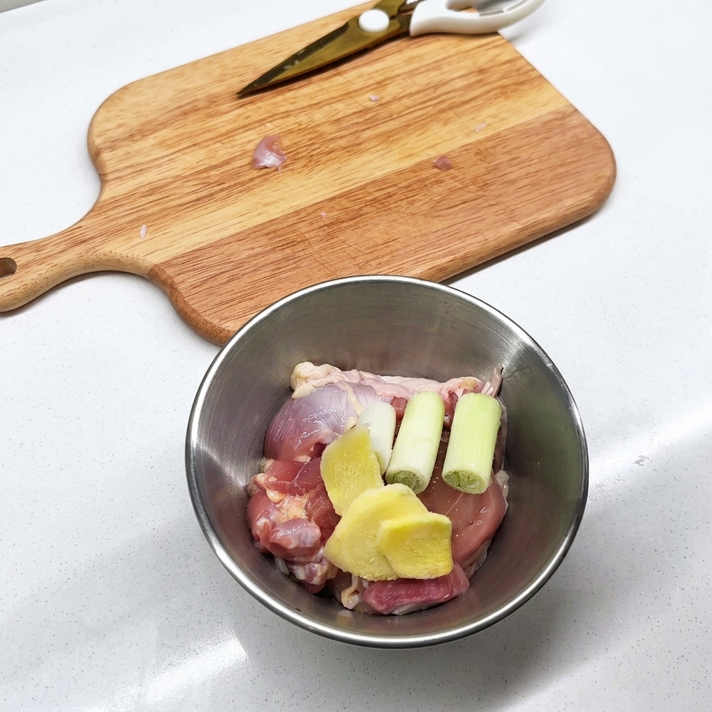 Tazón de metal con piezas de muslo de pollo crudo, rodajas de jengibre y tallos de cebolla verde.