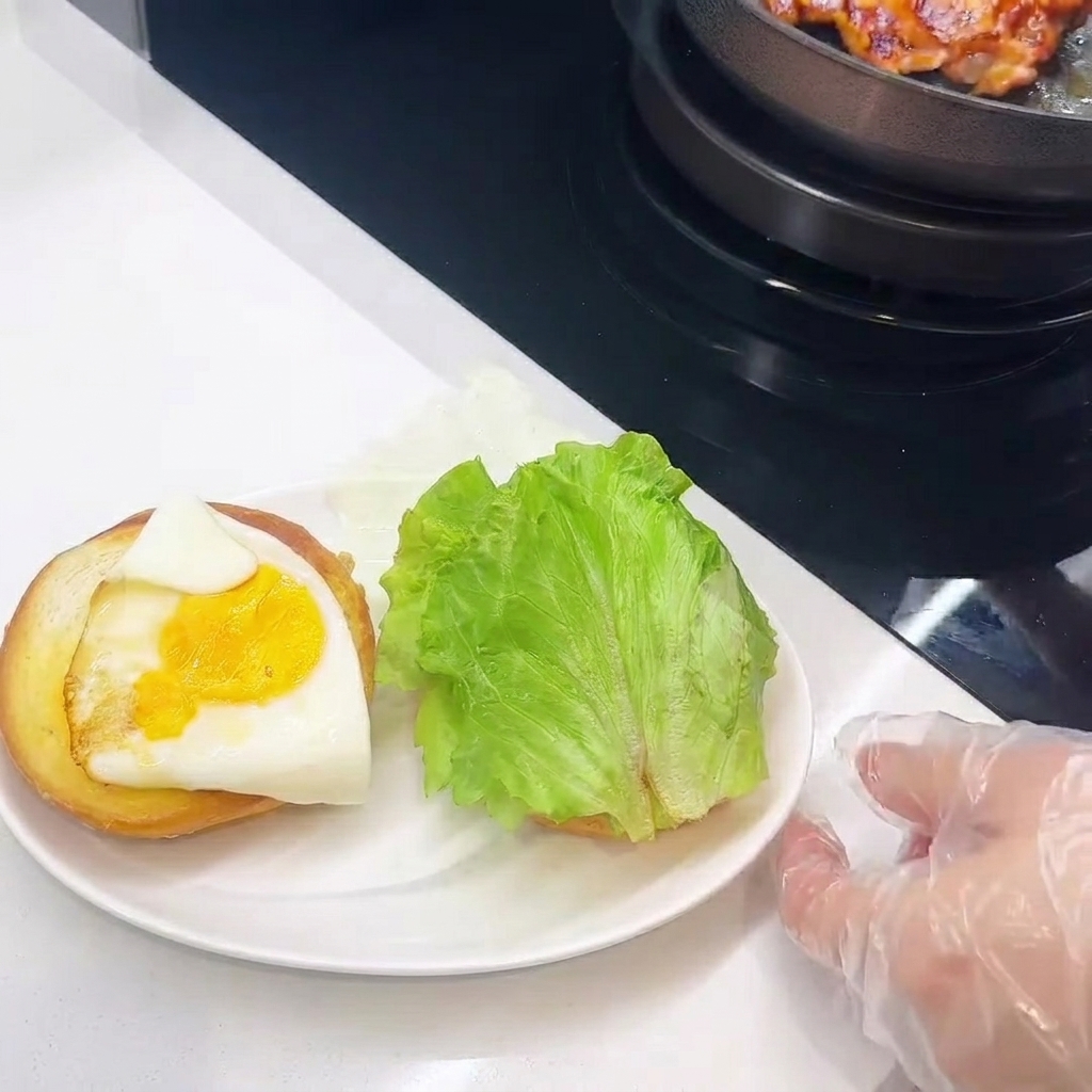 Vista de primer plano del armado de la hamburguesa, mostrando el pollo, queso, huevo y lechuga siendo apilados sobre el pan.