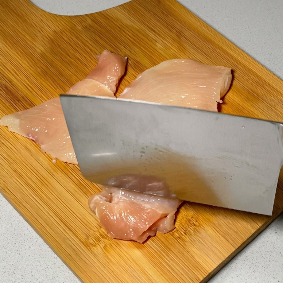 Un cuchillo de cocina utilizado para ablandar lonchas de pollo crudo sobre una superficie de madera.