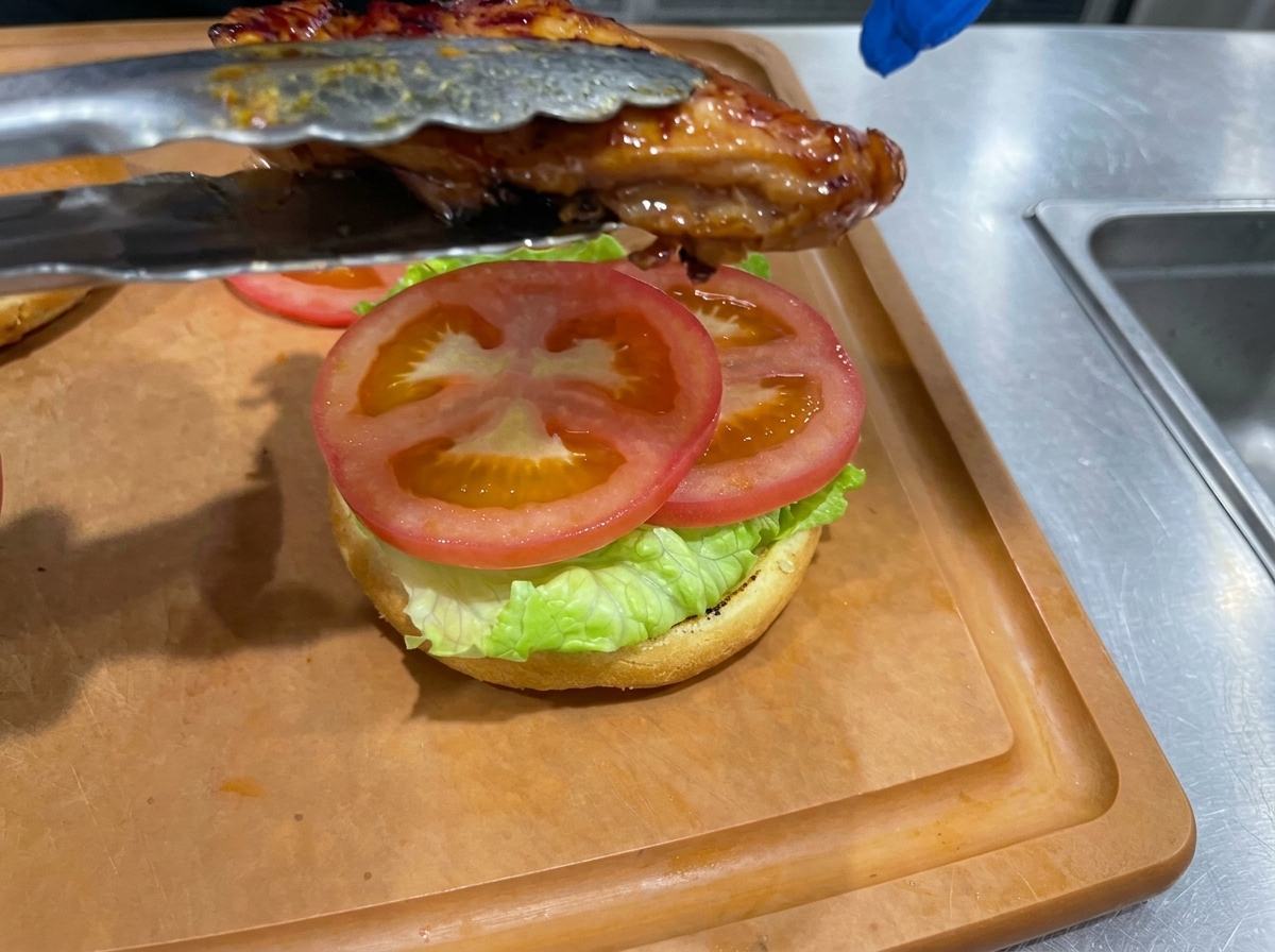 Un muslo de pollo teriyaki cocinado siendo colocado sobre la hamburguesa ya montada.