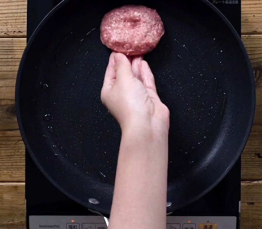 Una mano colocando una hamburguesa cruda en una sartén negra sobre una cocina de inducción.